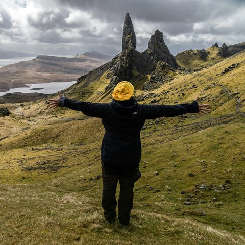 A man standing on a lush green hillside with arms spread in a yellow hat and black jacket