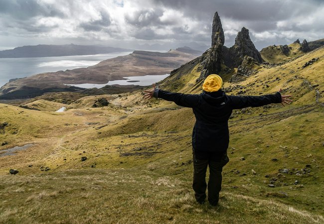 A man standing on a lush green hillside with arms spread in a yellow hat and black jacket