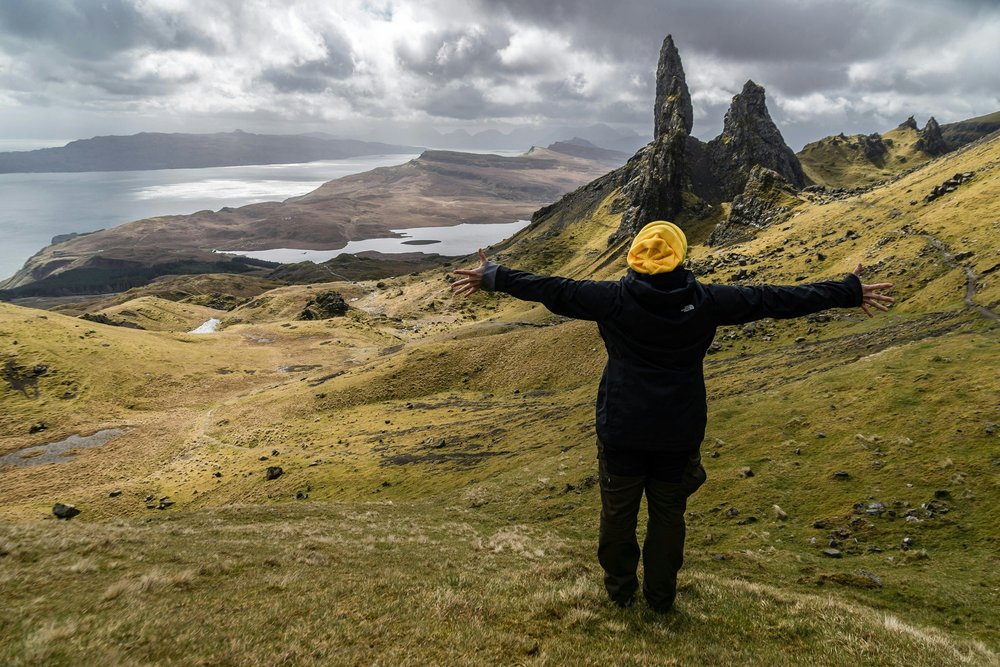 A man standing on a lush green hillside with arms spread in a yellow hat and black jacket