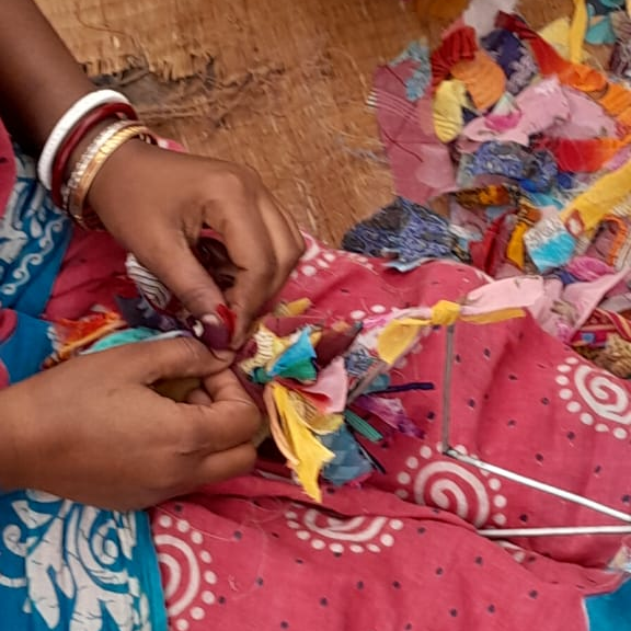 A close-up of a person’s hands tying together small, colourful fabric scraps, as part of a traditional craft or textile-making process. The person is wearing bangles and a bright patterned garment.