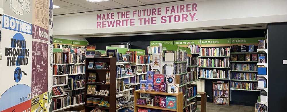 A bookstore interior with shelves filled with books, some display racks in the center, and wall signs above the shelves. A large message in pink letters reads "MAKE THE FUTURE FAIRER REWRITE THE STORY."