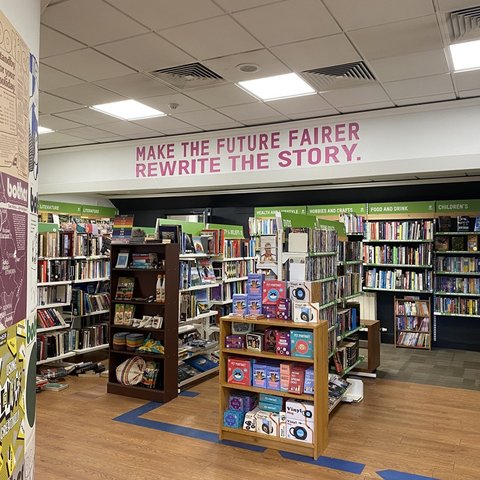 A bookstore interior with shelves filled with books, some display racks in the center, and wall signs above the shelves. A large message in pink letters reads "MAKE THE FUTURE FAIRER REWRITE THE STORY."