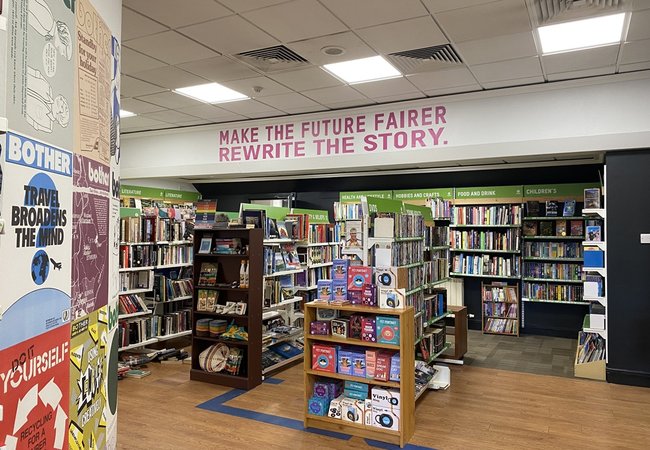 A bookstore interior with shelves filled with books, some display racks in the center, and wall signs above the shelves. A large message in pink letters reads "MAKE THE FUTURE FAIRER REWRITE THE STORY."