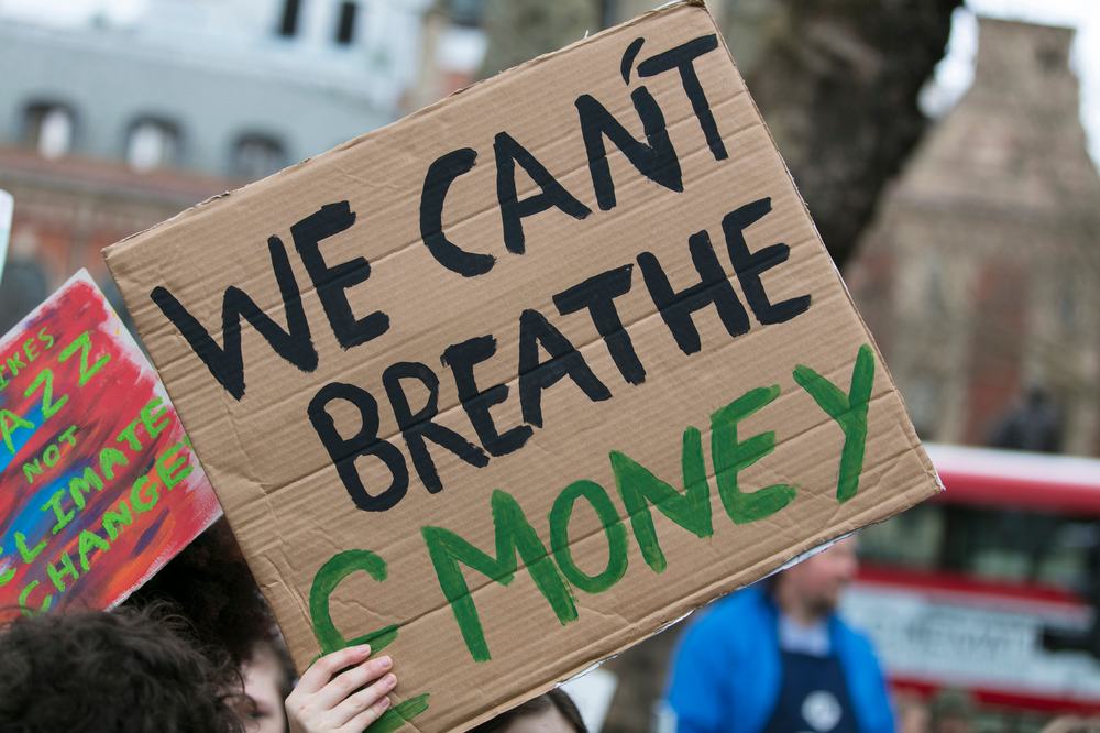 A climate protestor holds a sign that says 'we can't breathe money' there's a red double decker London bus in the background