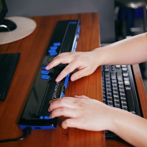 A person who is blind's hands on a braille technology assistive device in front of a computer.