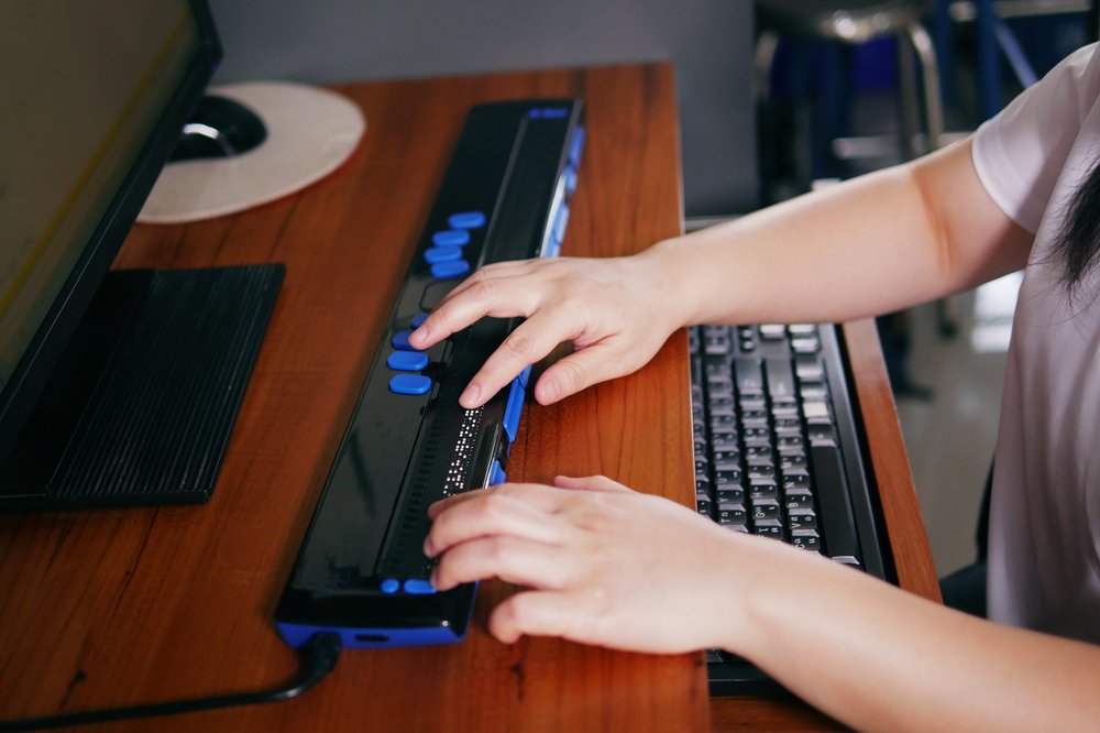 A person who is blind's hands on a braille technology assistive device in front of a computer.