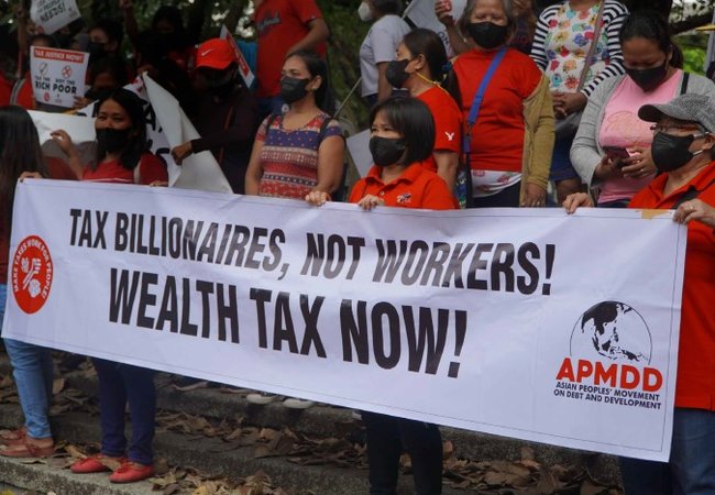 A group of women at a demonstration holding a large banner that reads "Tax billionaires, not workers! Wealth tax now!" The foreground sponsor logo is for the Asian Peoples' Movement on Debt and Development.