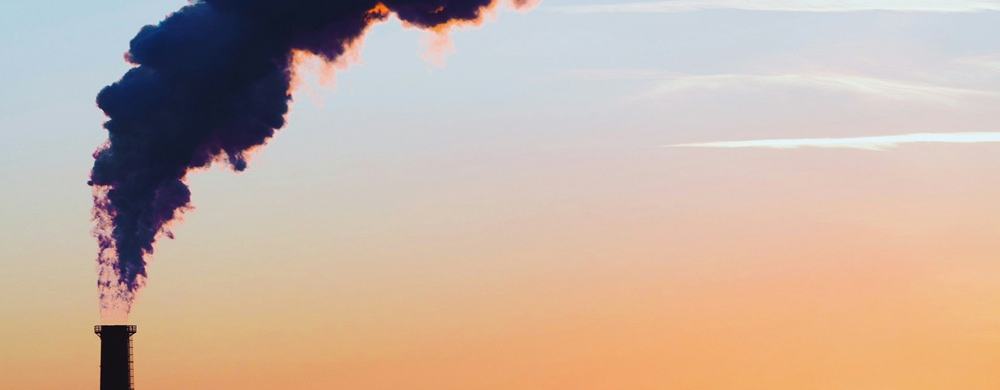 Chimney stack emitting a thick plume of dark smoke over a city skyline at sunset, with the bright sun low on the horizon and a plane flying in the distance.