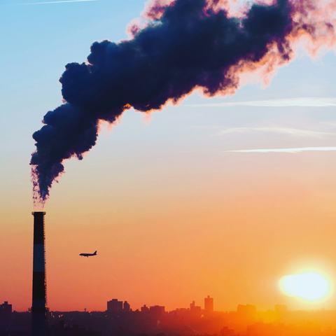 Chimney stack emitting a thick plume of dark smoke over a city skyline at sunset, with the bright sun low on the horizon and a plane flying in the distance.