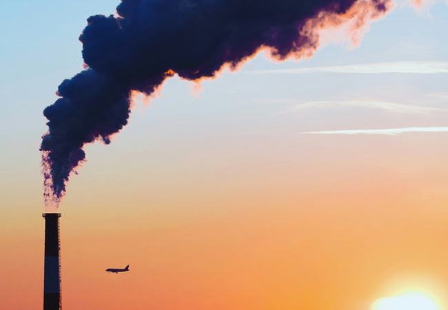 Chimney stack emitting a thick plume of dark smoke over a city skyline at sunset, with the bright sun low on the horizon and a plane flying in the distance.