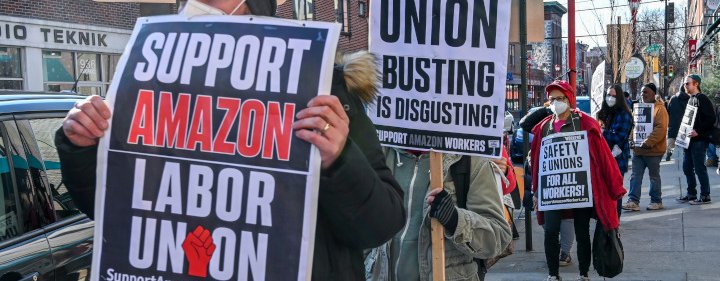 A group of men wearing coats, gloves and protective face masks on a city footpath holding a variety of placards that read: "Support Amazon labor union. supportamazonworkers.org", "Starbucks: union busting is disgusting" & "Safety & unions for all workers"