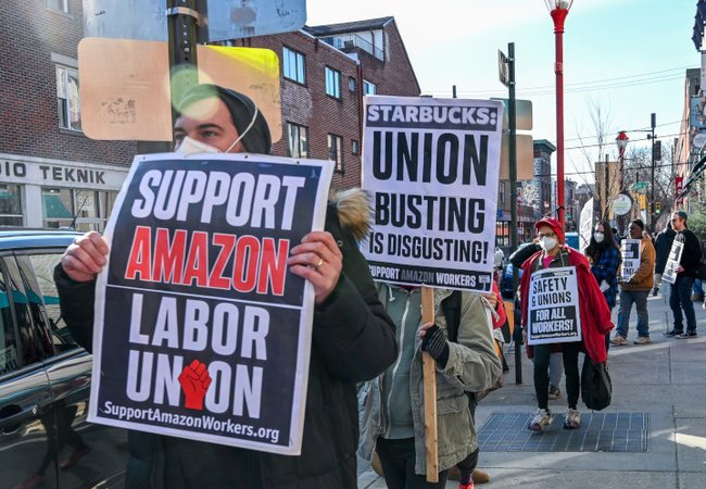 A group of men wearing coats, gloves and protective face masks on a city footpath holding a variety of placards that read: "Support Amazon labor union. supportamazonworkers.org", "Starbucks: union busting is disgusting" & "Safety & unions for all workers"