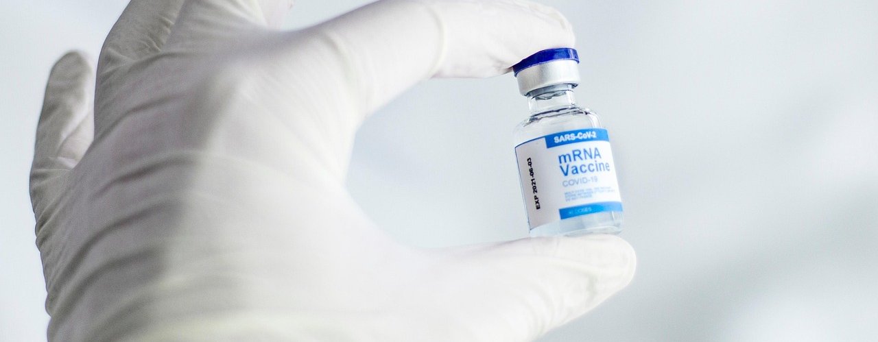 A hand inside a white surgical glove is holding a vial of the mRNA vaccine.