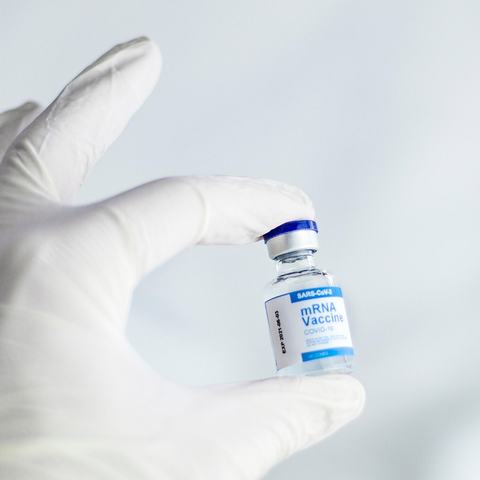 A hand inside a white surgical glove is holding a vial of the mRNA vaccine.