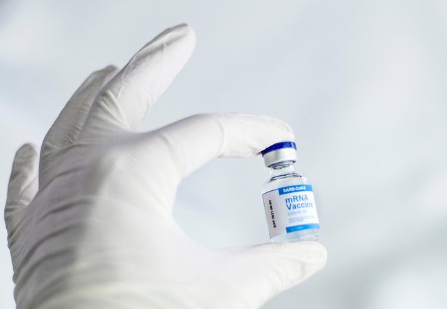 A hand inside a white surgical glove is holding a vial of the mRNA vaccine.