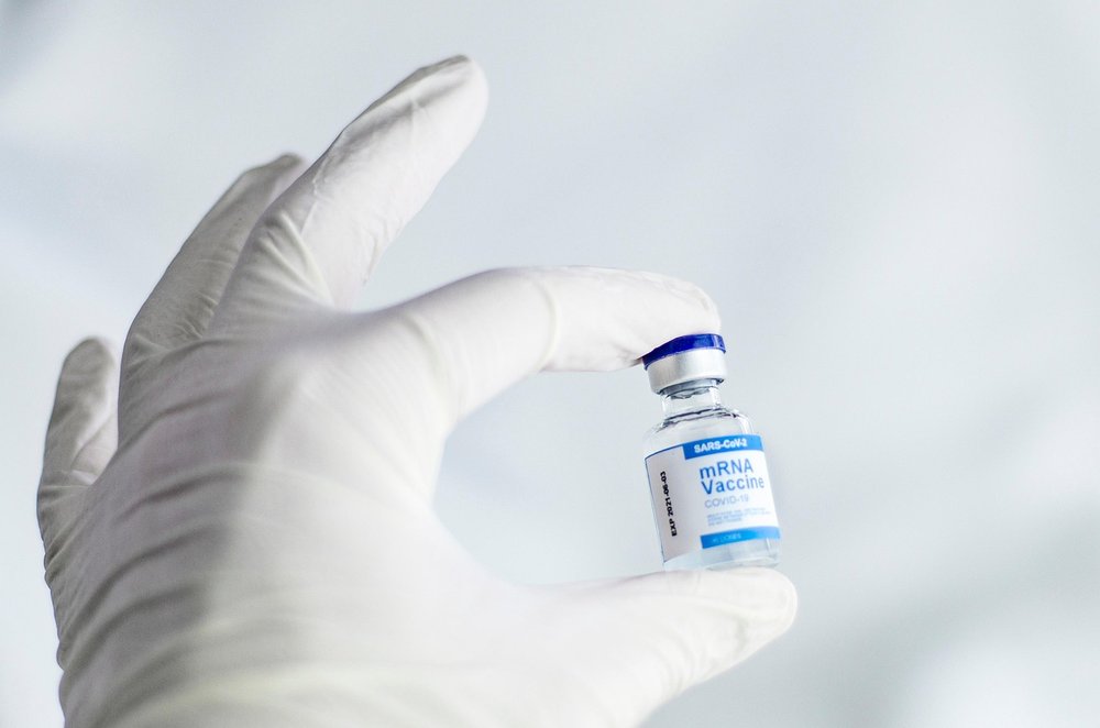 A hand inside a white surgical glove is holding a vial of the mRNA vaccine.