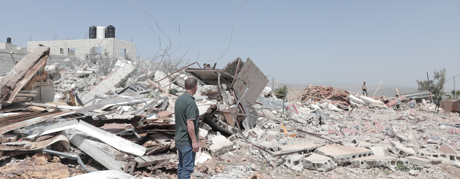 A man in the West Bank standing looking towards piles of rubble of a destroyed building. Other people can be seen looking at the rubble in the distance.