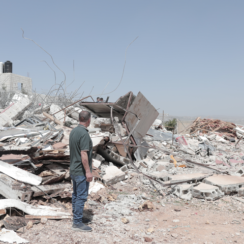 A man in the West Bank standing looking towards piles of rubble of a destroyed building. Other people can be seen looking at the rubble in the distance.
