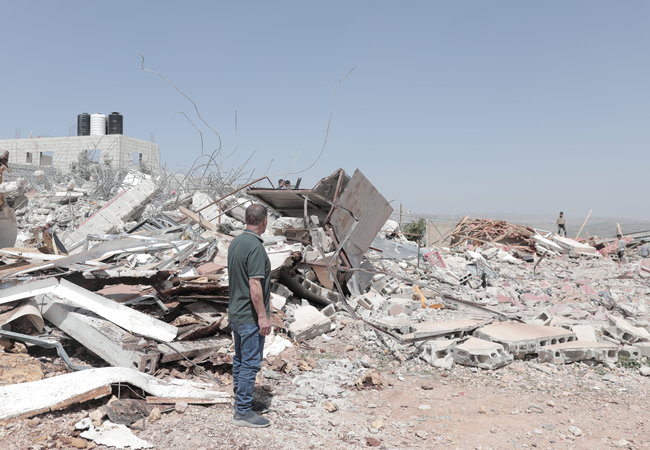 A man in the West Bank standing looking towards piles of rubble of a destroyed building. Other people can be seen looking at the rubble in the distance.