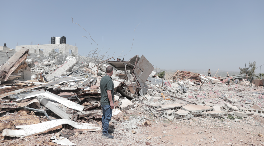 A man in the West Bank standing looking towards piles of rubble of a destroyed building. Other people can be seen looking at the rubble in the distance.