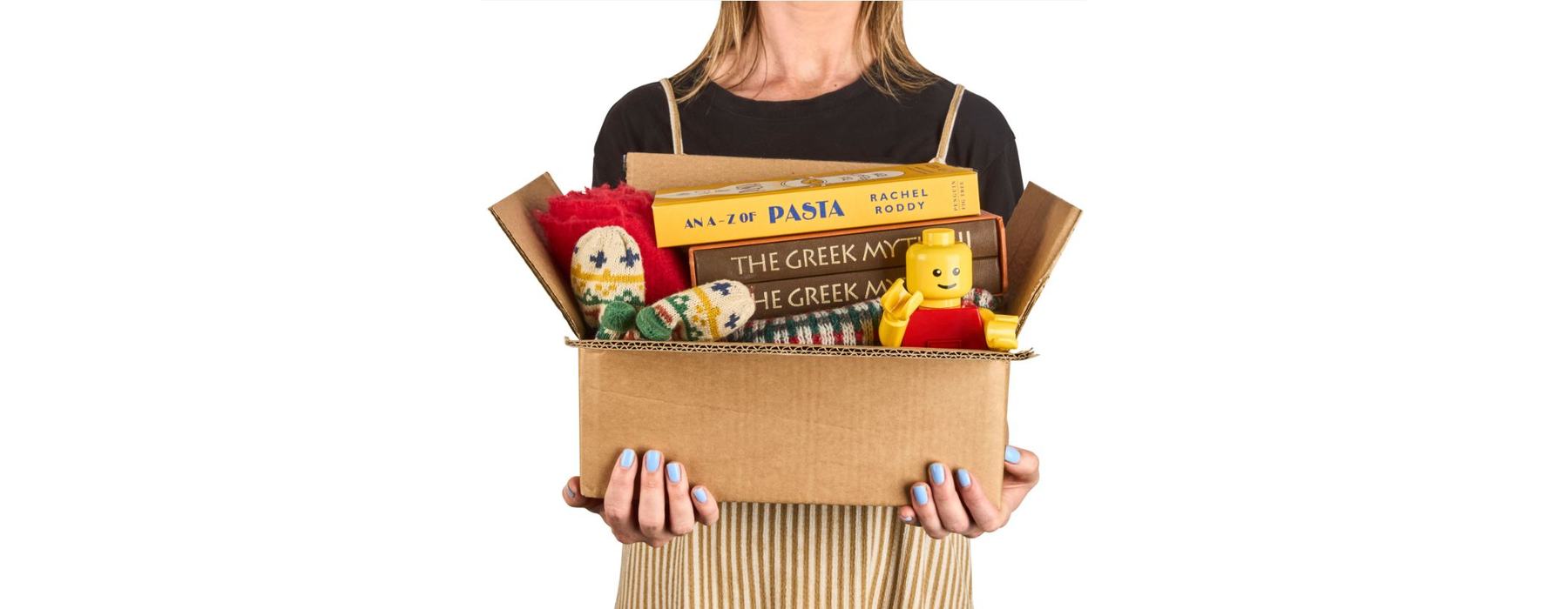 A woman is standing holding a cardboard box filled with donated charity items including books, a lego person and a knitted hat and gloves.