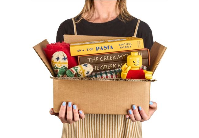 A woman is standing holding a cardboard box filled with donated charity items including books, a lego person and a knitted hat and gloves.