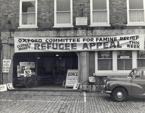 A black and white photo of a cobbled street with a large banner hanging over one building reading 'Oxford Committee for Famine Relief Refugee Appeal'