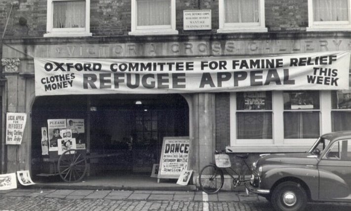 A black and white photo of a cobbled street with a large banner hanging over one building reading 'Oxford Committee for Famine Relief Refugee Appeal'